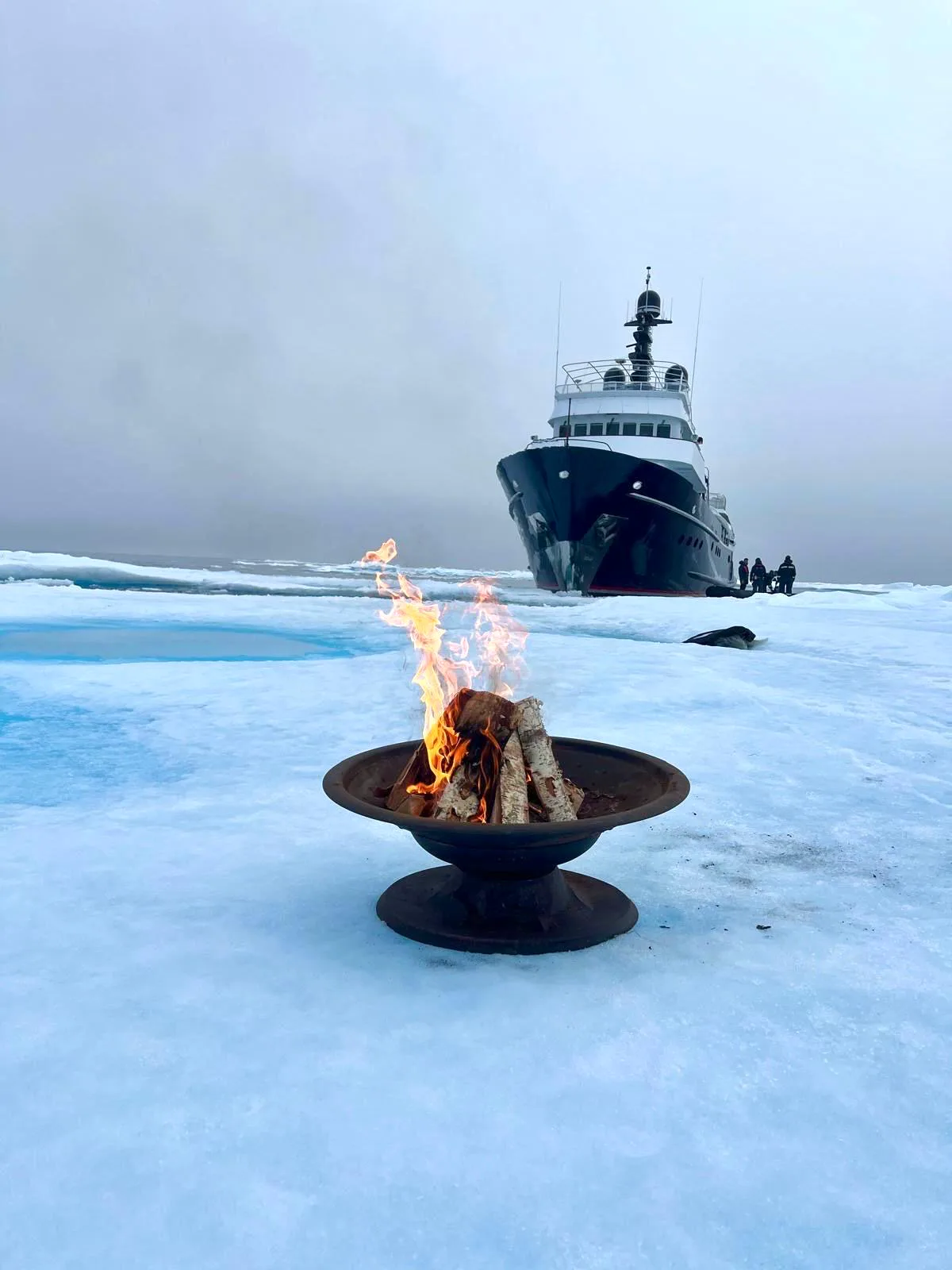 Fire bowl burning on pack ice with the expedition yacht anchored behind, Svalbard